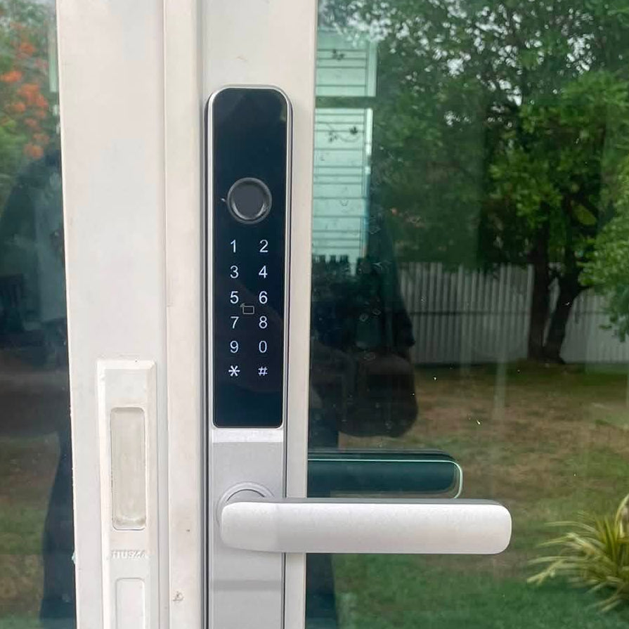 Smart door lock with black keypad panel and silver lever fitted to a white framed glass door, garden behind