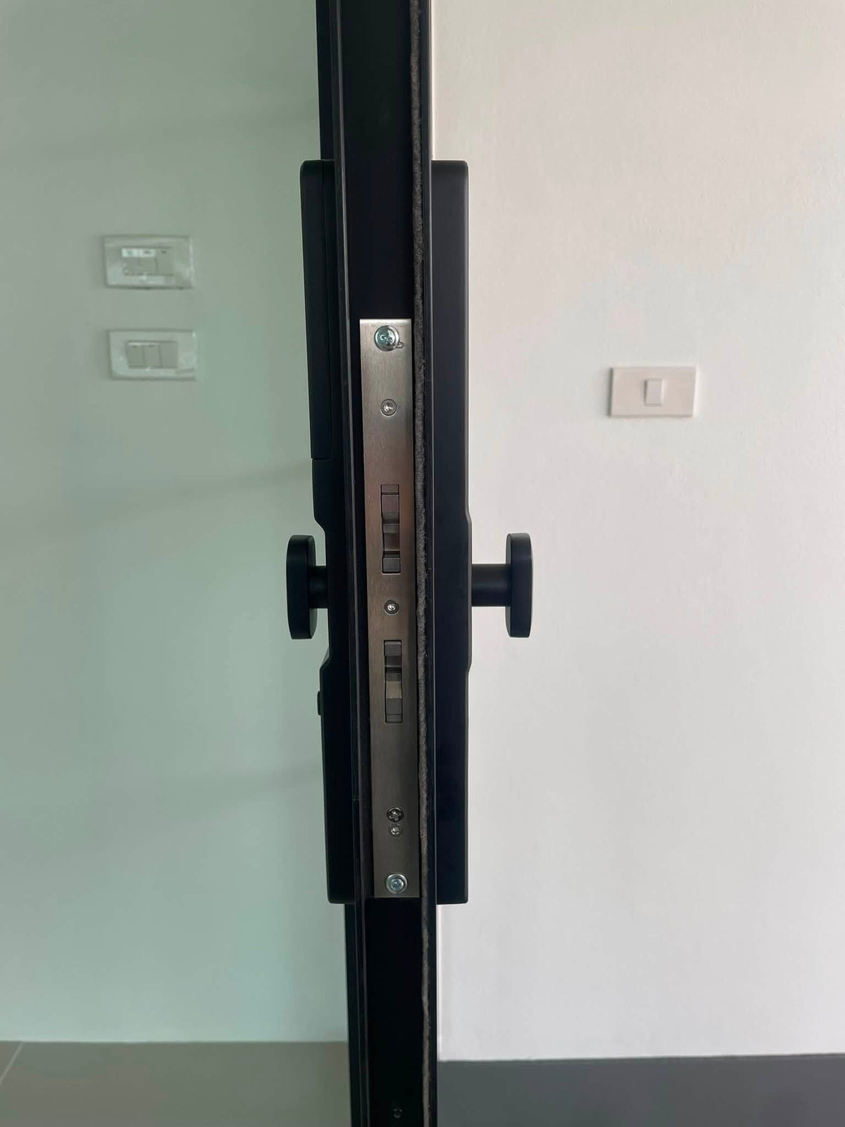Black short-handle smart door lock on a white interior door, light switches on the wall beside it